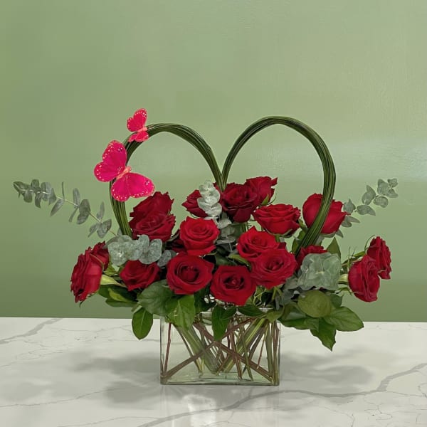 Red roses arranged in a glass vase with a heart-shaped frame and pink butterfly accents.