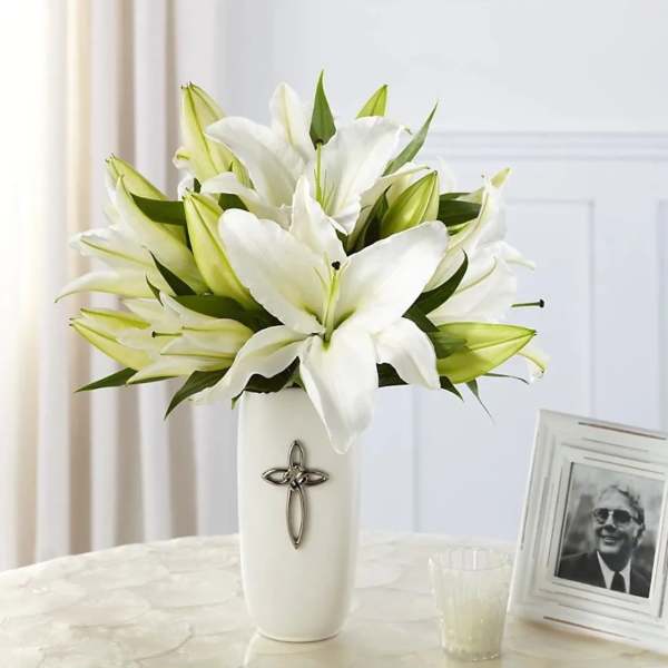White lilies arranged in a tall white vase with a cross emblem