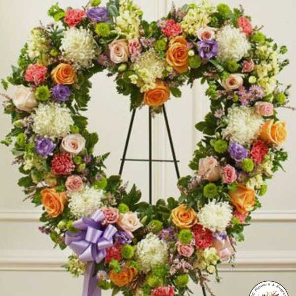 Heart-shaped standing wreath of mixed roses, carnations, and mums in soft colors with a lavender bow