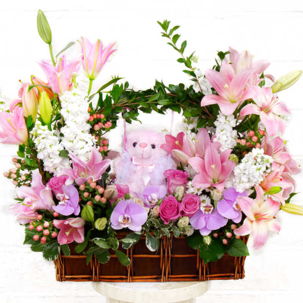 Pink lilies and orchids arranged in a wicker basket with a teddy bear