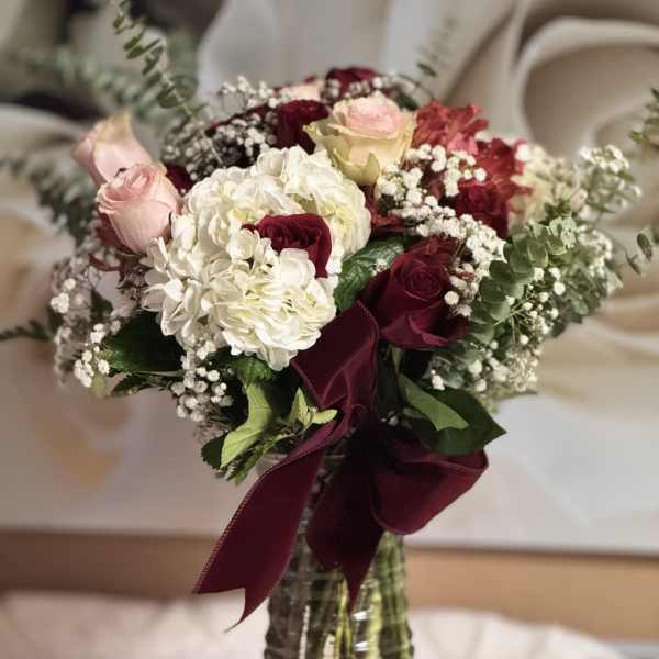 Tall vase arrangement of red and pink roses with white hydrangeas and baby's breath, tied with a burgundy bow.