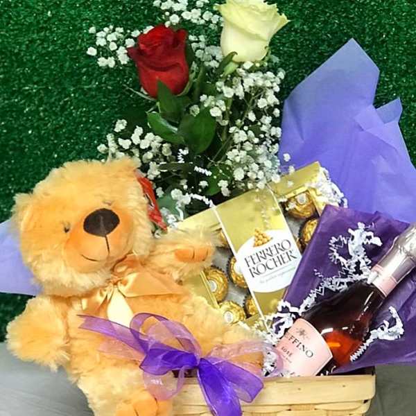 Gift basket with teddy bear, red and white roses, chocolates, and a pink sparkling drink wrapped with purple ribbon
