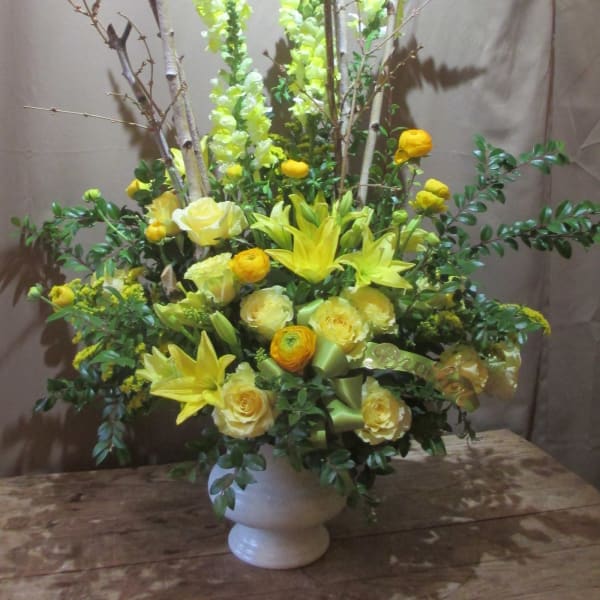 Tall yellow flower arrangement with lilies, roses, and branches in a white urn vase