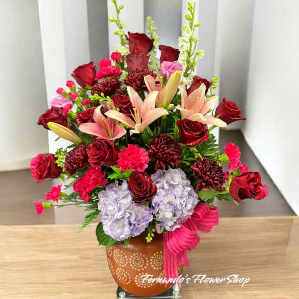 Large mixed arrangement of red roses, pink lilies, bright carnations, and lavender hydrangeas in an orange pot
