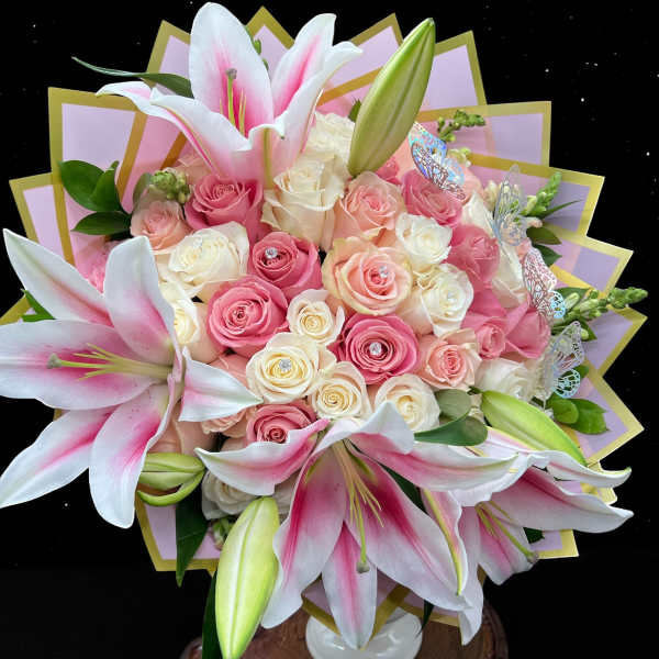 Pink lilies and cream and pink roses arranged in a white vase with decorative wrap and butterfly accents