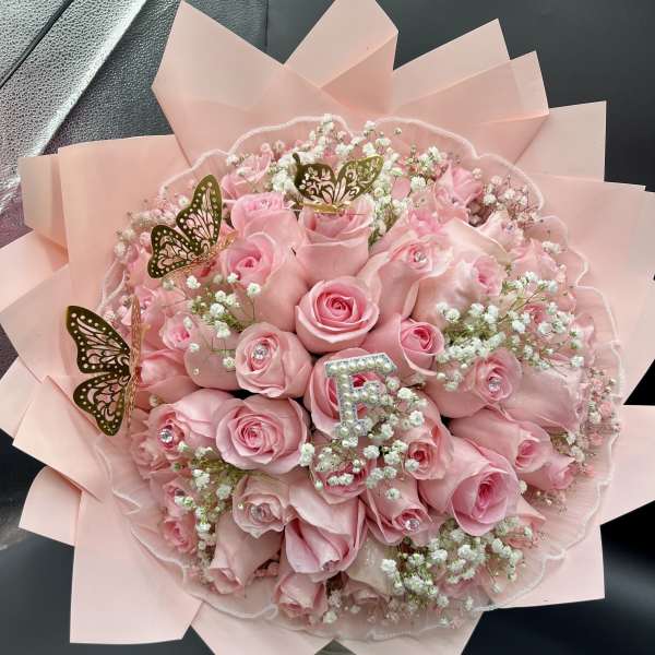 Round bouquet of pink roses and white filler flowers with gold butterfly accents wrapped in pale pink paper.
