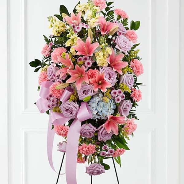 Standing spray of pink lilies, lavender roses, and carnations on an easel with lavender ribbon.
