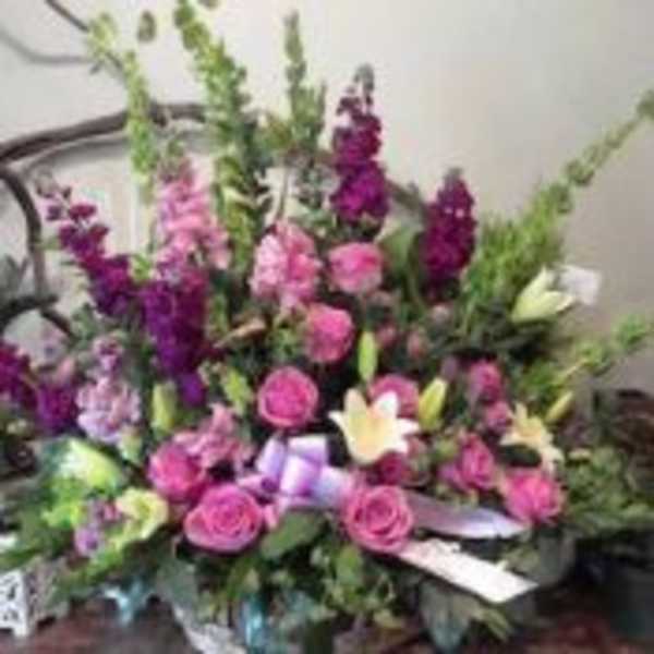 Tall arrangement of pink roses, purple flowers, and pale lilies in a container with a lavender bow.