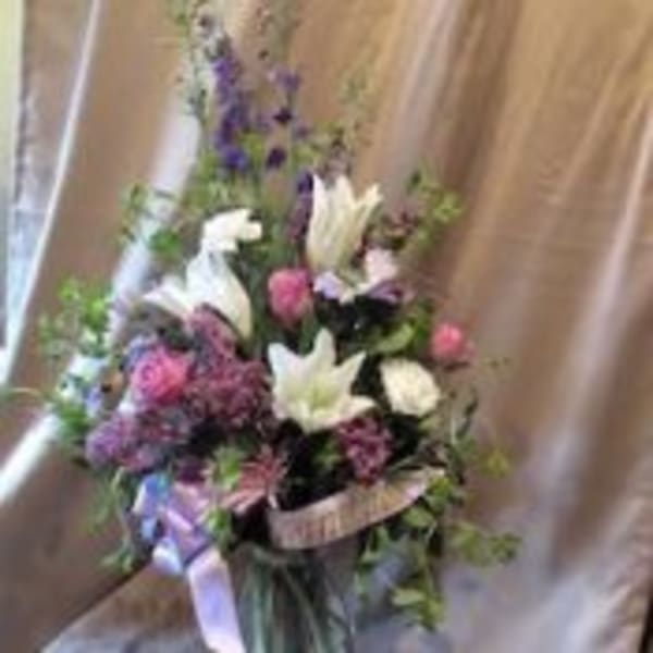 Tall mixed arrangement of white lilies, pink roses, and purple flowers in a clear glass vase with ribbons
