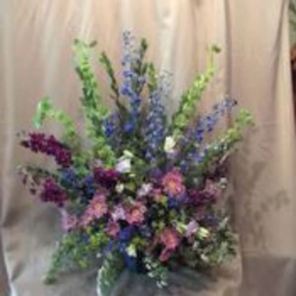 Tall fan-shaped arrangement of purple, blue, pink, and green flowers against a neutral draped background