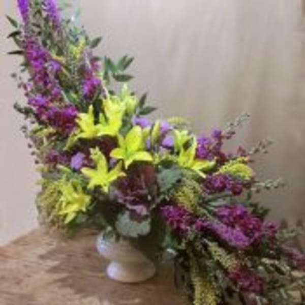 Tall asymmetrical arrangement of yellow lilies and purple flowers in a white pedestal vase