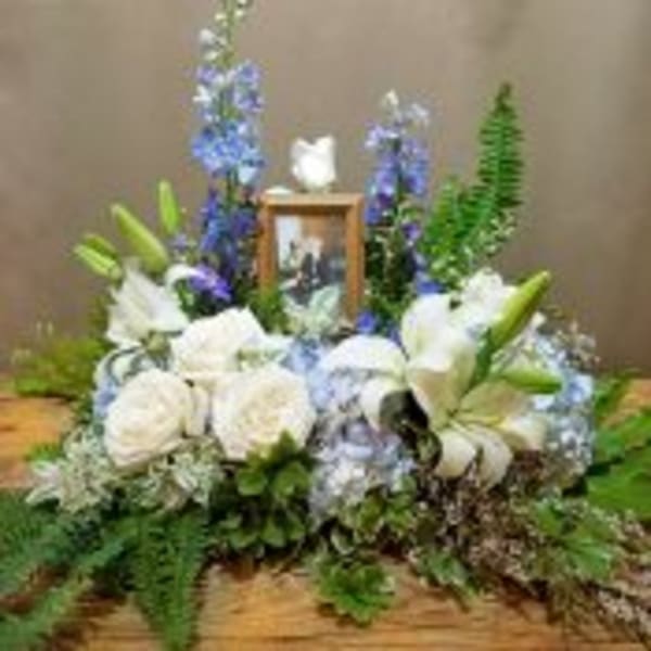 Sympathy floral spray of white roses and lilies with blue flowers around a framed photo and dove figurine