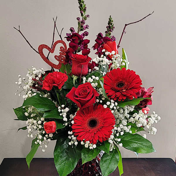 Red roses, gerbera daisies, carnations, and baby’s breath in a red glass vase with a glitter heart pick.