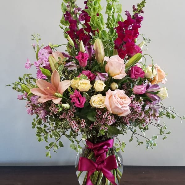 Tall mixed bouquet of pink, peach, and yellow flowers with green spikes in a clear vase with a magenta bow