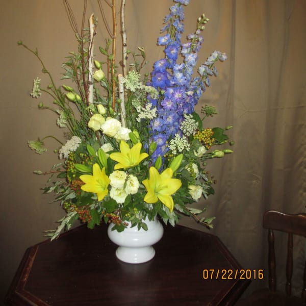 Tall arrangement of yellow lilies, white blooms, and blue delphinium in a white vase with branches