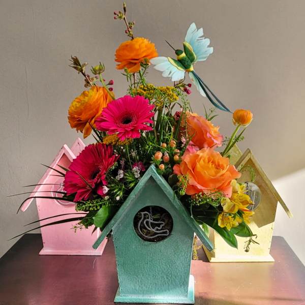 Bright mixed bouquet in a teal birdhouse container with orange, pink, and yellow flowers and a decorative bird pick