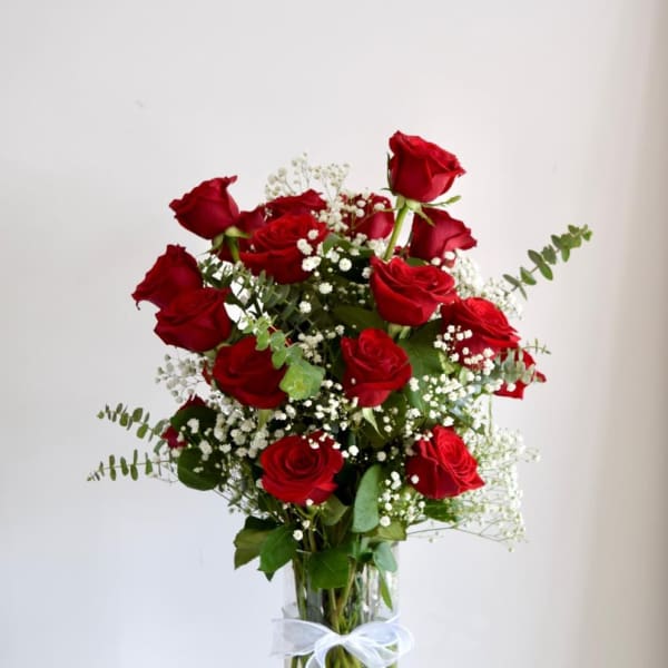 Tall arrangement of long-stem red roses with small white filler flowers in a clear vase with a white bow.