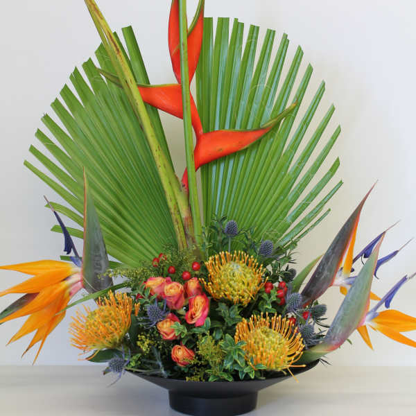Tall tropical arrangement with orange and yellow flowers in a black shallow bowl.