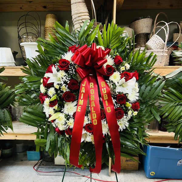 Standing sympathy wreath with red roses, white flowers, and red ribbon on a metal easel.