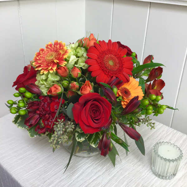 Low round arrangement of red roses, gerbera daisies and mixed orange blooms in a clear glass bowl vase