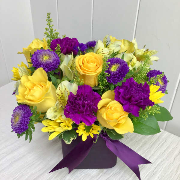 Low cube arrangement of yellow roses and daisies with purple carnations and asters in a purple vase with ribbon