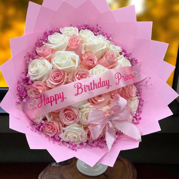 Round bouquet of pink and white roses with pink wrapping, birthday ribbon, and bow on a small pedestal stand