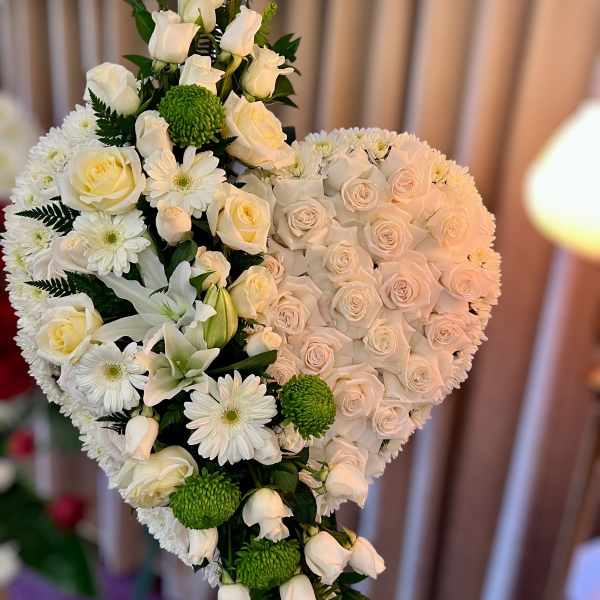 Standing heart-shaped spray of white roses, daisies, and lilies with green accents on an easel.