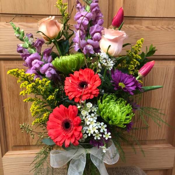 Mixed bouquet with pink roses, red gerbera daisies, green mums, and tulips in a glass vase with ribbon