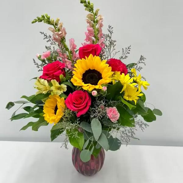 Mixed bouquet with sunflowers, hot pink roses, snapdragons, and daisies in a red glass vase