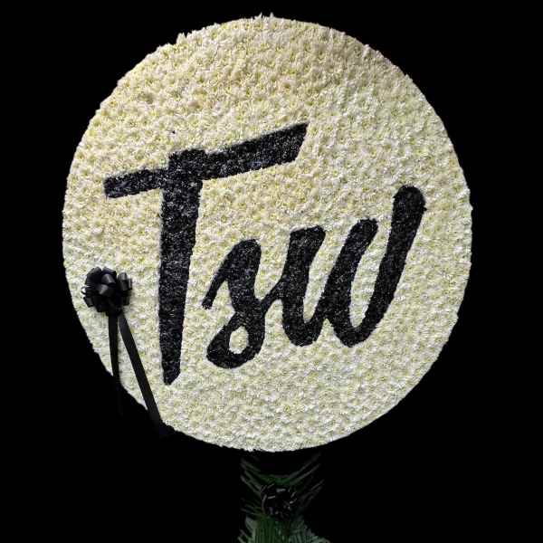 Round white floral tribute with black lettering and ribbon, mounted above a small floral base