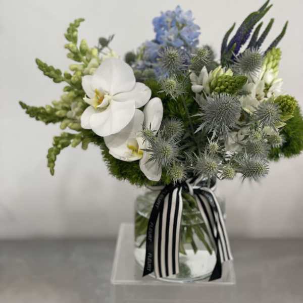 Low glass vase arrangement with white orchids, blue flowers, and green accents tied with a striped ribbon.