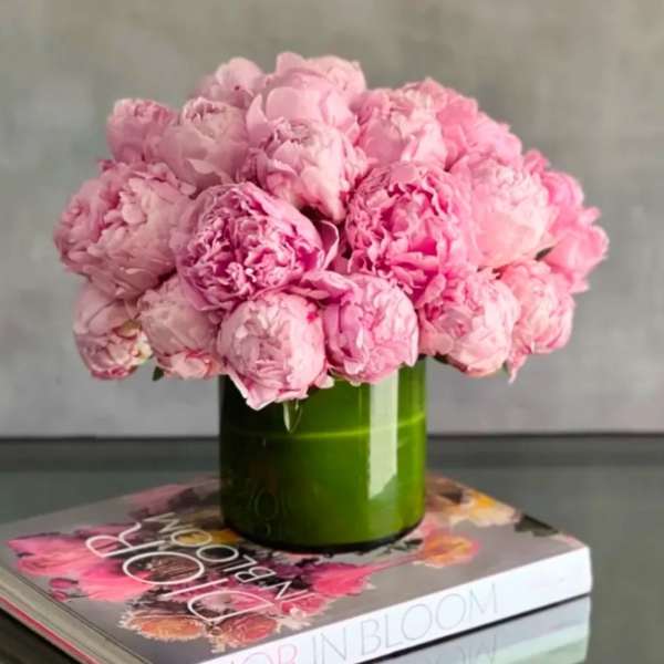 Tight cluster of pink peonies in a green cylinder vase set on a floral coffee table book