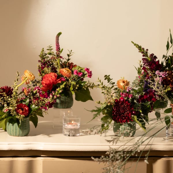 Three floral arrangements in green vases on a table with candles and scissors