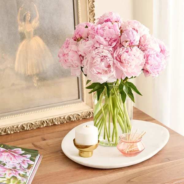 Pink peonies in a clear glass vase on a tray with a candle and small dish