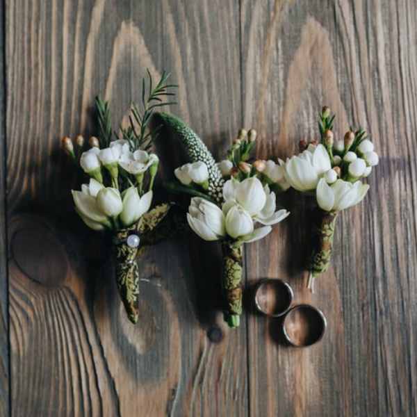 Three white floral boutonnieres on a wooden surface with two rings
