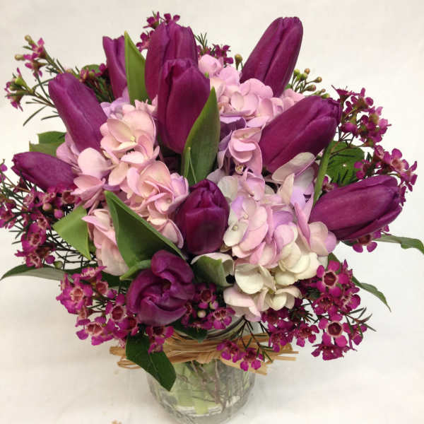Purple tulips with pink hydrangeas and small blooms in a clear glass jar vase tied with raffia.