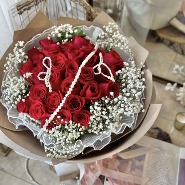 Hand-tied bouquet of red roses and white filler flowers wrapped in paper with draped pearl strands.