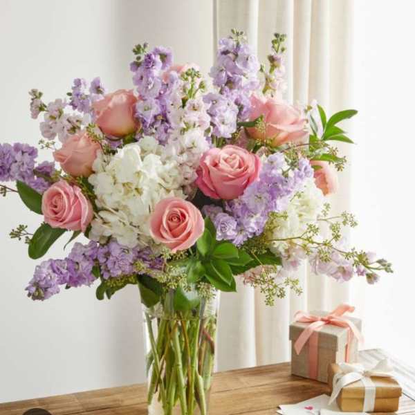 Pink roses and lavender flowers in a clear glass vase beside wrapped gifts