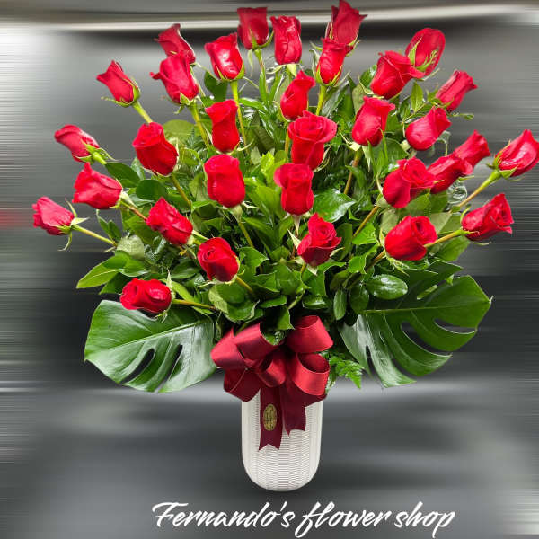 Tall arrangement of red roses in a white vase with large glossy leaves and a red ribbon bow
