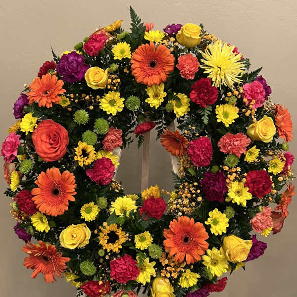 Bright circular wreath of orange gerbera daisies, yellow roses, and mixed flowers on a wooden easel.