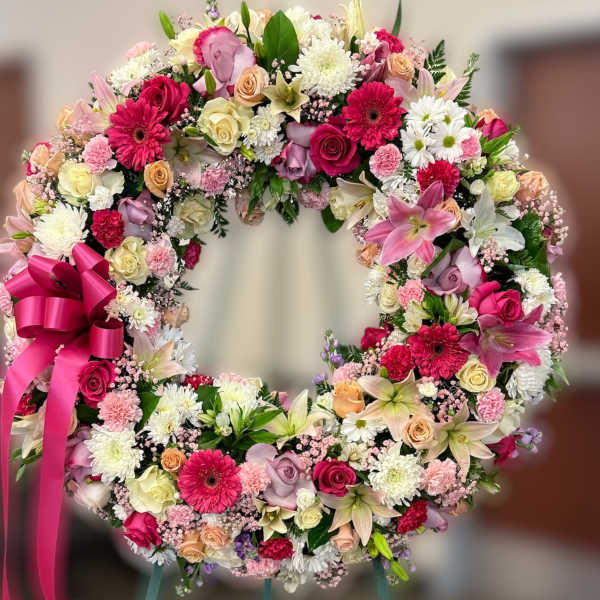 Standing wreath of pink, white, and peach flowers with a bright pink ribbon bow on an easel.