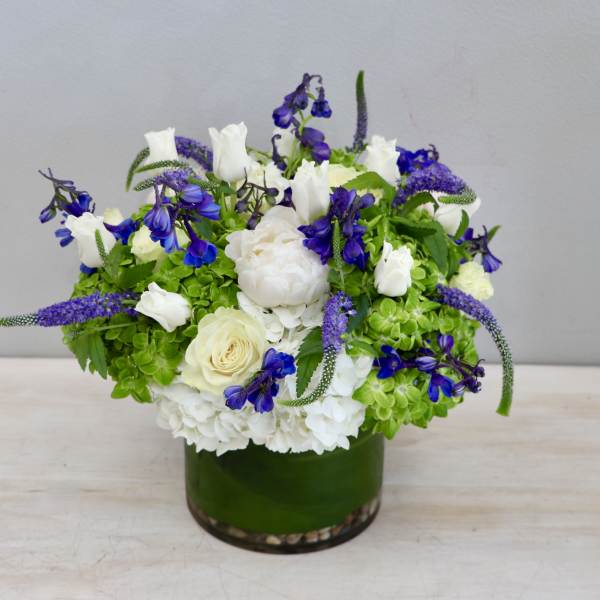 Low round arrangement of white and blue flowers with green accents in a glass vase wrapped with a green leaf.