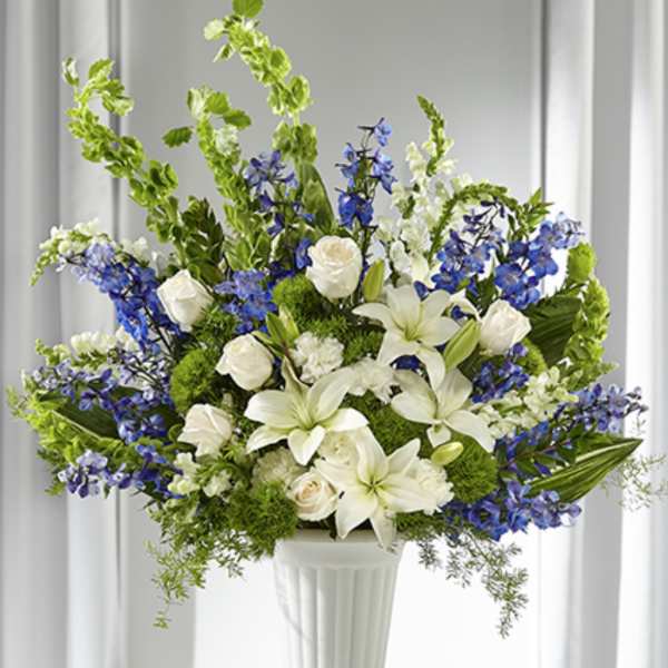 Tall white and blue arrangement with lilies, roses, delphinium and Bells of Ireland in a white pedestal vase.