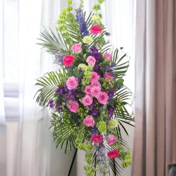 Tall standing spray with pink roses, purple flowers, and green accents on an easel.