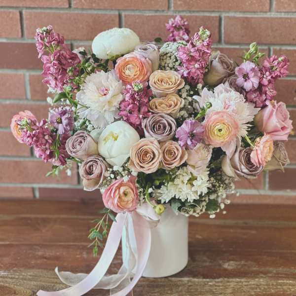 Pastel arrangement of roses, ranunculus, and peonies in a white vase with ribbon