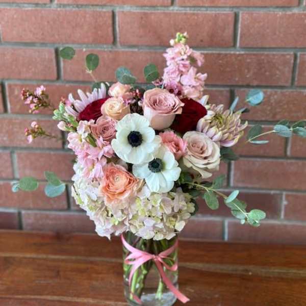 Tall mixed bouquet of soft pink, white, and peach flowers in a clear vase with a pink ribbon.