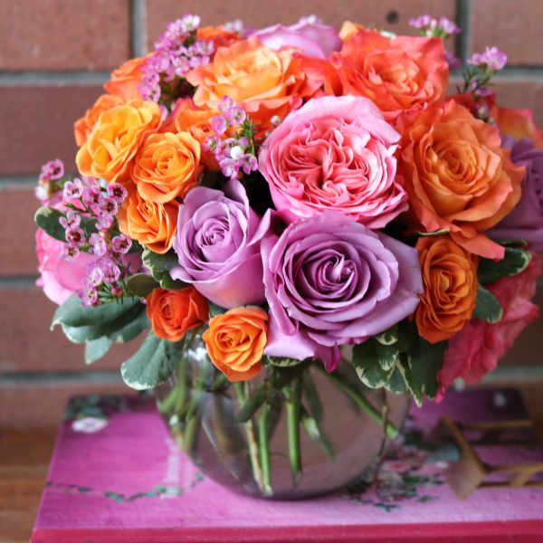 Round arrangement of orange, pink, and lavender roses in a clear glass bowl vase