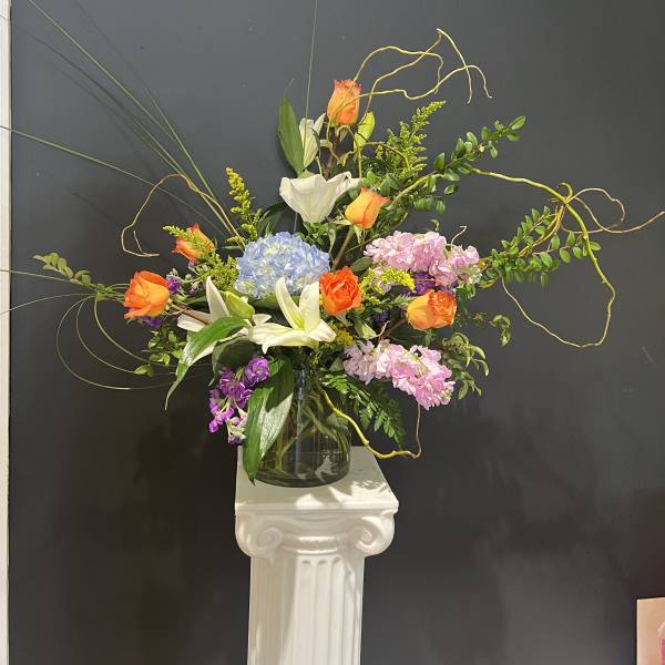 Tall mixed flower arrangement in a glass vase with orange roses, lilies, and hydrangea