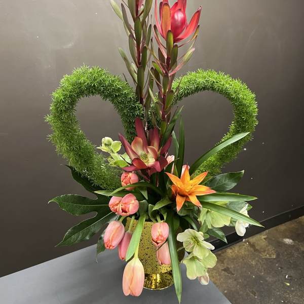 Tall floral arrangement with pink and orange blooms in a gold vase, framed by green heart shapes.