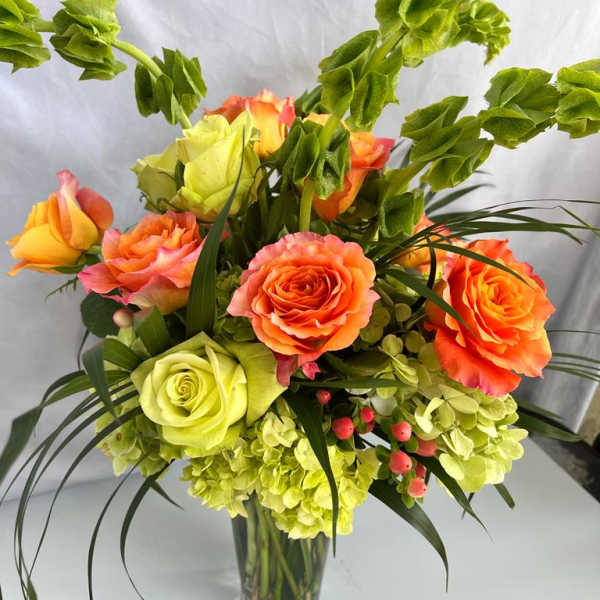 Tall arrangement of orange and green roses, green hydrangeas, and bells of Ireland in a clear glass vase